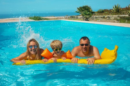 Foto de Una familia alegre de tres, madre, padre e hijo disfrutando de un día de piscina soleado salpicando en una balsa inflable de color amarillo brillante durante las vacaciones de verano - Imagen libre de derechos