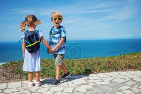 Foto de Rubio chico y chica con mochilas cogidas de la mano mirando al mar desde la colina, niño gritando, mira a la cámara en el caluroso día de verano en Portugal - Imagen libre de derechos