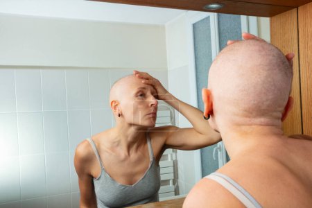 Photo for Pensive hairless woman ill of breast cancer in a bathroom, looking at herself in the mirror while touching her bald head - Royalty Free Image