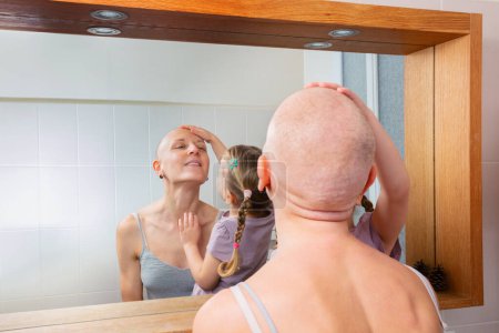 Téléchargez les photos : Une mère chauve souriant tandis que sa fille touchait sa tête, regardant leur reflet dans un miroir de salle de bain, moment intime de la personne aux prises avec le cancer - en image libre de droit