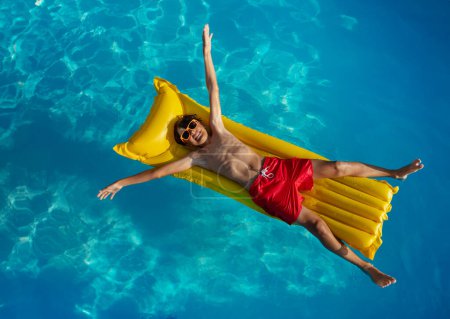 Foto de Un niño descansando en una carroza amarilla en una piscina azul cristalina, con gafas de sol y pantalones cortos rojos - Imagen libre de derechos