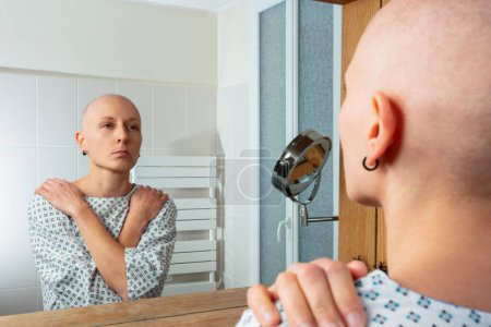 Foto de Persona sin pelo en prendas de salud mira pensativamente en su reflejo en el espejo de un baño - Imagen libre de derechos