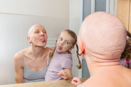 Photo for A young child and an adult woman without hair fighting cancer playfully sticking out their tongues in front of a mirror - Royalty Free Image
