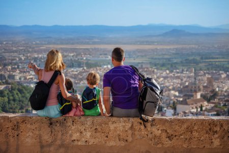Photo for Family of travelers sit on viewpoint wall show to kids Alhambra ancient palace on their summer vacations in Granada, Spain - Royalty Free Image