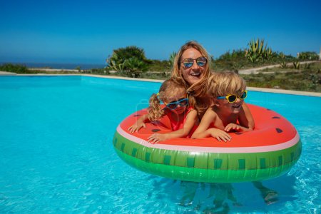 Foto de Mujer con gafas de sol, sonriendo y abrazando a dos niños más pequeños nadan en sandía inflable - Imagen libre de derechos