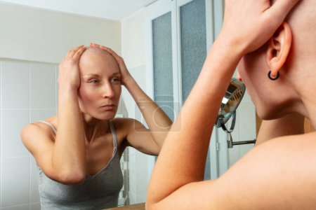 Photo for A thoughtful bald woman after cancer treatment is caught in a moment of reflection as she touches her head and looks into the bathroom mirror - Royalty Free Image