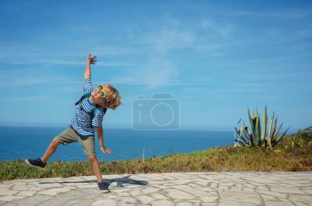 Foto de Niño rubio feliz en traje de verano con gafas de sol bailando sobre el fondo del mar entusiasmado con las vacaciones en Portugal - Imagen libre de derechos