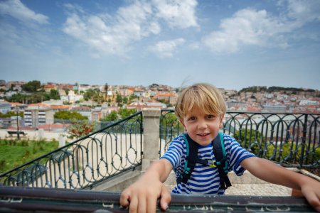 Foto de Niño rubio sonriente en el mirador disfruta de las impresionantes vistas de Lisboa sosteniendo balaustrada con mochila - Imagen libre de derechos