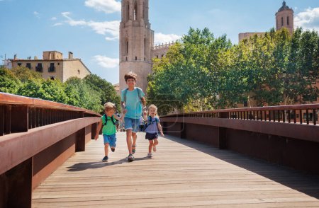 Foto de Tres niños viajeros con mochilas cruzando el puente Sant Feliu en el centro de Girona en su tour turístico de verano en España - Imagen libre de derechos