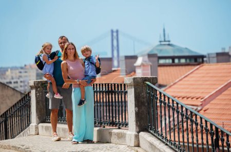 Mom and dad two kids on family vacations in Portugal standing looking at camera with famous Lisbon bridge on background in summer day