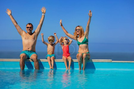 Téléchargez les photos : Deux adultes et leurs enfants s'amusent à plonger les pieds dans une piscine, avec l'océan derrière, un vaste ciel bleu au-dessus d'eux. - en image libre de droit