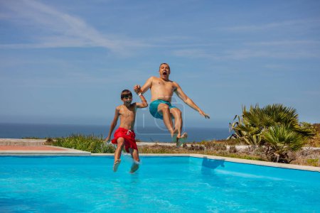 Téléchargez les photos : Saut ludique dans une piscine d'eau par un homme et un jeune avec un vaste océan au loin, plaisir pendant les vacances d'été - en image libre de droit