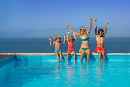 Téléchargez les photos : Heureux famille lever les mains dans le geste d'excitation et pendre leurs pieds dans les eaux d'une piscine avec la mer fusionnant dans le ciel devant - en image libre de droit