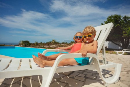 Foto de Lindo niño y niña, dos hermanos descansando en tumbonas blancas disfrutar del sol de verano, relajarse en sillas de la piscina con la zona de baño del complejo en el fondo - Imagen libre de derechos