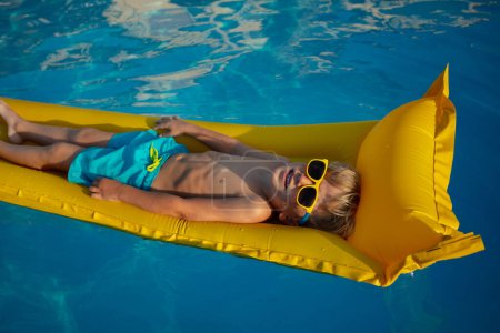 Téléchargez les photos : Un jeune enfant se prélasse sur un radeau gonflable jaune dans une piscine, portant lunettes de soleil et maillots de bain bleus - en image libre de droit