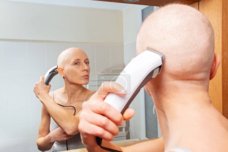 Foto de Mujer lucha contra el cáncer afeitándose la cabeza con un zumbador, concentrándose en la imagen del espejo - Imagen libre de derechos