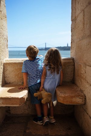 Foto de Dos niños pequeños turistas miran por la ventana de Torre de Belem en Lisboa en su gira de visita a Portugal, ver el famoso puente 25 Abril - Imagen libre de derechos
