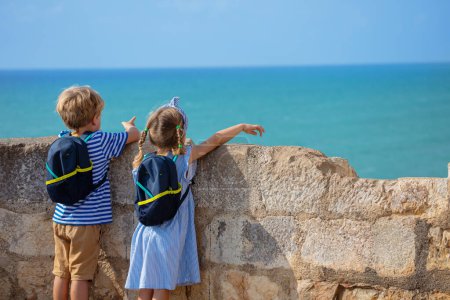 Foto de Jóvenes turistas niño y niña con pequeñas mochilas apuntan al mar desde las paredes del castillo Peniscola, disfrutar de vacaciones europeas de verano - Imagen libre de derechos