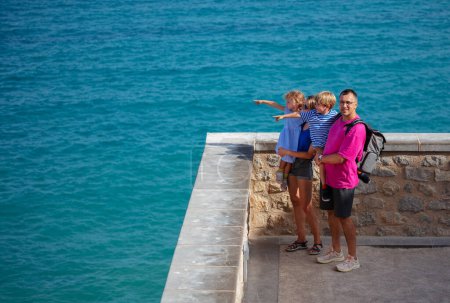 Foto de Parada familiar turística de verano en el mirador del castillo Peniscola apuntando con los dedos al mar turquesa y sonriendo - Imagen libre de derechos