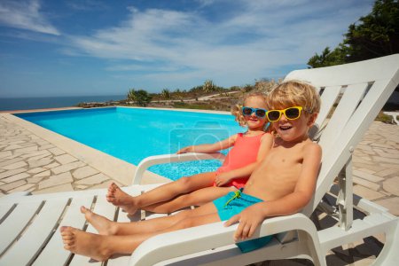 Foto de Un niño y una niña descansando en tumbonas blancas junto a una piscina serena, con gafas de sol de colores, bajo un cielo azul durante las vacaciones de verano - Imagen libre de derechos
