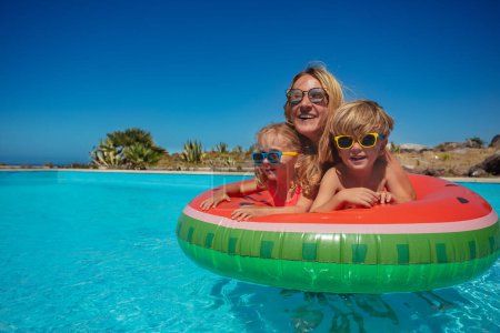 Foto de Una mujer y dos niños pequeños en gafas de sol flotando en un anillo inflable de sandía en una piscina brillante bajo cielos azules claros - Imagen libre de derechos