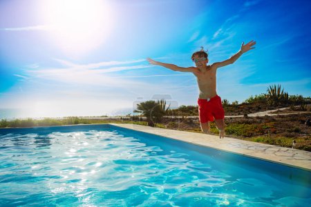 Foto de Un niño en pantalones cortos rojos salta jubilosamente junto a una piscina brillante en un día soleado - Imagen libre de derechos
