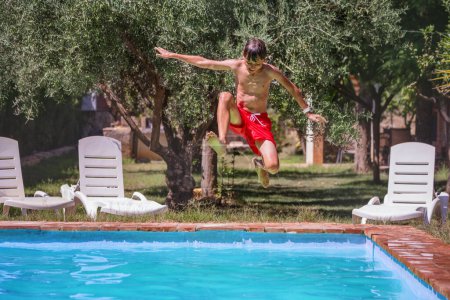 Téléchargez les photos : Adolescent mignon porter rouge baignade secondes loin de plonger dans les eaux bleues invitantes d'une piscine, sur fond de fauteuils roulants blancs et feuillage vert. - en image libre de droit