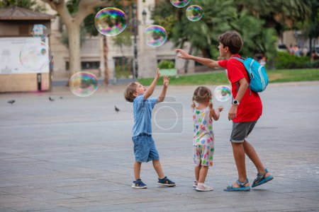 Foto de Grupo de niños juegan y tratan de atrapar las enormes burbujas de jabón en el día de verano en la plaza de la ciudad en Valencia, España - Imagen libre de derechos