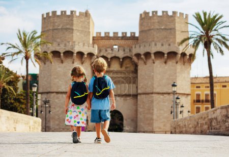 Foto de Dos niños viajeros caminan juntos en la calurosa Valencia frente a las famosas Torres de Serranos durante sus vacaciones de verano en España, vista desde atrás - Imagen libre de derechos