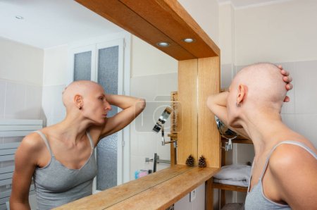 Photo for A pensive bald female individual fight with cancer, examines her reflection in a mirror within a luminous washroom - Royalty Free Image