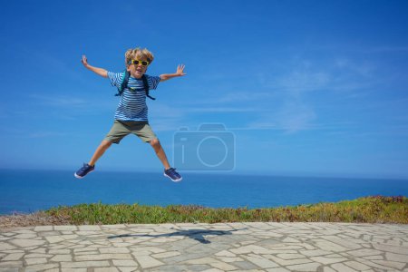 Téléchargez les photos : Enfant blond heureux sauter haut sur fond de mer portant une tenue marine et des lunettes de soleil, excité de voir l'océan - en image libre de droit