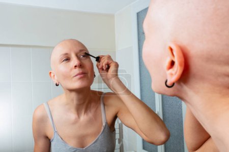Téléchargez les photos : Femme positive combattant le cancer s'engageant dans le rituel de beauté de l'application de mascara dans une salle de bain à la maison - en image libre de droit