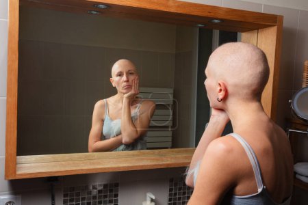 Foto de Una mujer sin pelo enferma de cáncer observa su propia imagen en el espejo del baño, colocando mano sobre cara de una manera pensativa - Imagen libre de derechos
