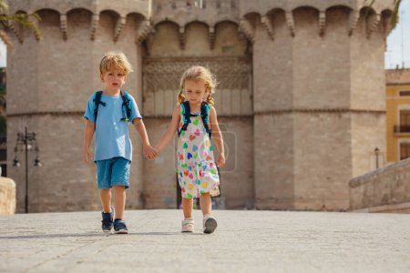 Photo for Two children hold hands, walk in sunny Valencia in front of famous Torres de Serranos during their summer trip in Spain - Royalty Free Image