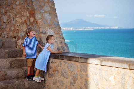 Foto de Dos hermanos, niño y niña de vacaciones se paran en la pared de Peniscola mirando al hermoso mar Mediterráneo, sonríen, disfrutan de su estancia en España - Imagen libre de derechos