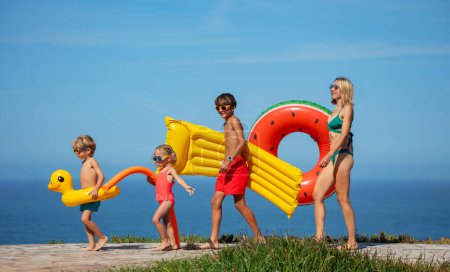 Téléchargez les photos : Belle famille, mère et trois enfants se préparent pour une journée de natation, portant des gonflables lumineux, avec une vue panoramique sur l'océan sur la toile de fond - en image libre de droit