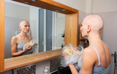 Foto de Mujer vulnerable mira en el espejo, cuidadosamente agarrando un peluquín, reflexionando sobre su calvicie - Imagen libre de derechos