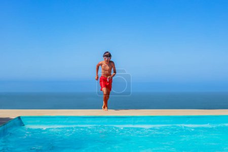 Foto de Adolescente niño usar pantalones rojos con gafas capturadas en movimiento y a punto de tomar una inmersión en la piscina cristalina contra un horizonte marino - Imagen libre de derechos