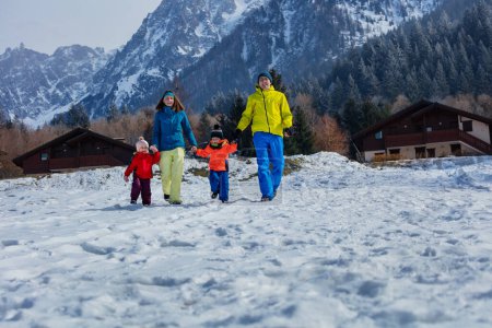 Foto de Familia feliz con niños corriendo juntos tomados de la mano en la nieve blanca en el pueblo francés nevado de los Alpes y las montañas en el fondo vistiendo traje de invierno - Imagen libre de derechos