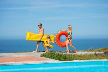 Foto de Niño, sus padres equipados para las actividades de la piscina pasan con anillos inflables y flotadores, el horizonte del mar a la vista - Imagen libre de derechos