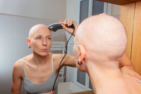 Foto de Una mujer calva se está cortando la cabeza con unas tijeras eléctricas mientras se mira al espejo - Imagen libre de derechos