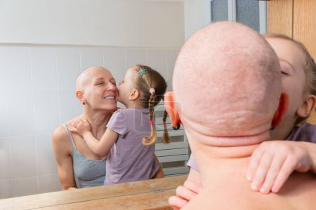 Téléchargez les photos : Dans une salle de bain bien éclairée, un enfant picore affectueusement la joue d'une femme souriante sans cheveux qui lutte contre le cancer - en image libre de droit