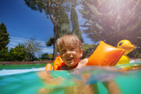 A cheerful kid sporting orange arm rings plays in a turquoise swimming pool on a sunny day, with an inflatable duck in the background