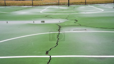 Empty outdoor green basketball court with visible cracks and white linesThe court awaits players on what appears to be an overcast day, with no equipment or people in sight.