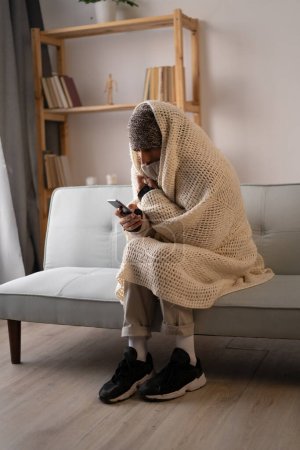 Young man dressed in winter clothes feeling cold sitting at home with no heating, gaming on smartphone. No heating concept.