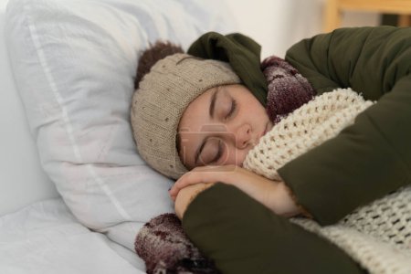 Téléchargez les photos : Dormir jeune femme dans une maison froide et non chauffée en hiver dans des vêtements chauds. Pauvres gens brisés incapables de payer pour l'électricité. La vie après la guerre. Pas de chaleur, concept de crise énergétique - en image libre de droit