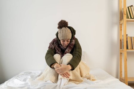 Photo for Angry young woman warmly dressed in a cold house sits on the bed feeling cold inside the house. Copy space - Royalty Free Image