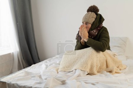 Traurige Weibchen sitzen bei eisiger Kälte in warmer Mütze auf dem Bett und zittern vor Kälte. Unzufriedene junge Frauen fühlen sich zu Hause schlecht und leiden unter Heizungsproblemen. Zu Hause vor der Kälte frieren