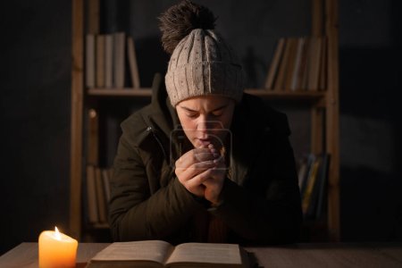 Freezing young woman in winter clothes warms her hands on lights with candles. Shutdown of heating and electricity, power outage, blackout, load shedding or energy crisis. copy space.