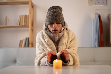 Müder Mann, der zu Hause bei Kerzenschein die Hände wärmt, ohne Heizung. Abschaltung von Heizung und Strom, Stromausfall, Stromausfall, Lastentlastung oder Energiekrise